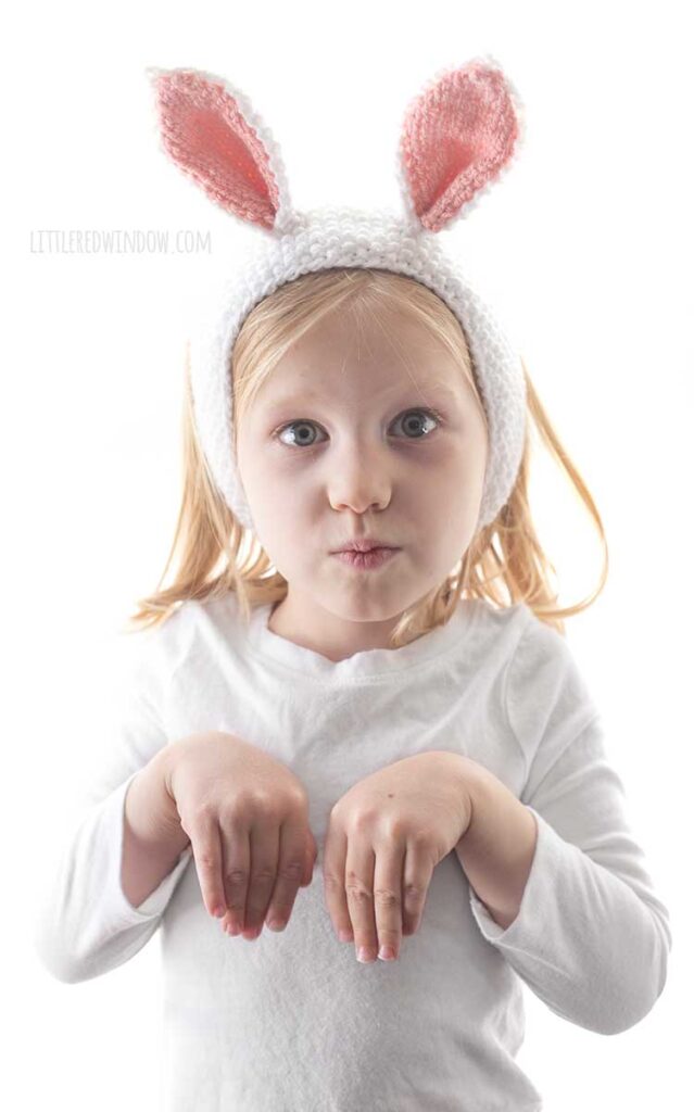 little girl with hands up pretending to be a bunny while wearing white seed stitch knit headband with white and pink bunny ears on top
