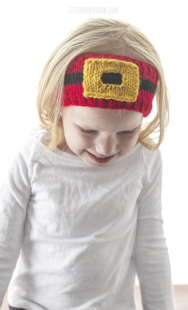 little girl in white shirt looking down and wearing a red knit headband with a black stripe and gold santa belt buckle on it