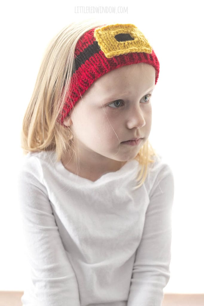 little girl in white shirt looking off to the right and wearing a red knit headband with a black stripe and gold santa belt buckle on it