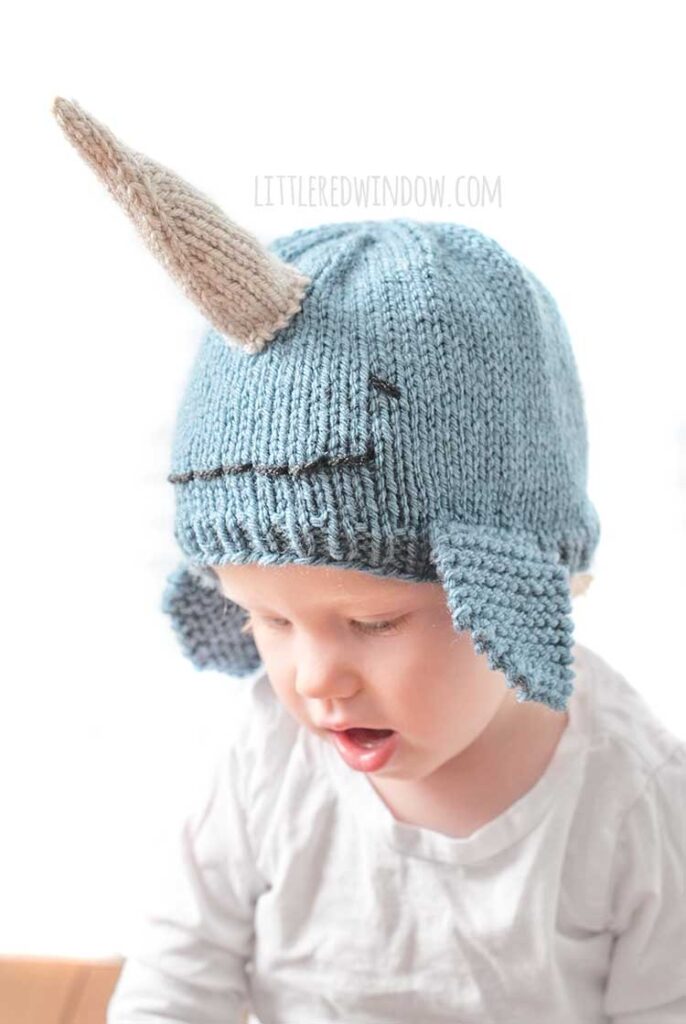 child in white shirt wearing a medium blue knit hat that looks like a narwhal in front of a white background looking down at their lap