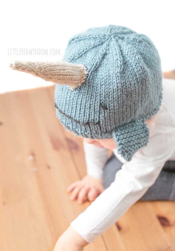 child in white shirt wearing a medium blue knit hat that looks like a narwhal crawling on a wood surface