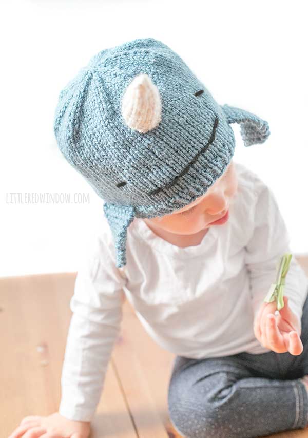 child in white shirt wearing a medium blue knit hat that looks like a narwhal in front of a white background looking at something in their hand