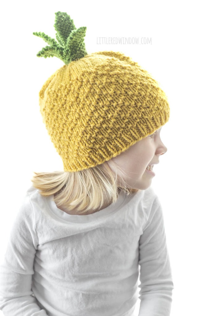 girl in white shirt wearing yellow knit pineapple hat with green leaves on top looking off to the right