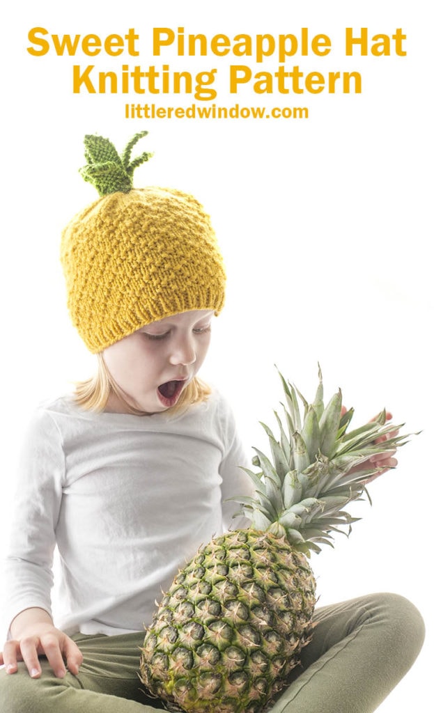 little girl looking down with surprise at a pineapple in her lap while wearing a pineapple knit hat