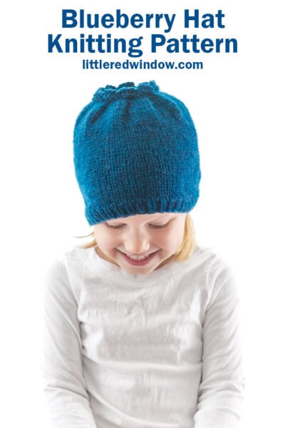 little girl in white shirt looking down and wearing a blue knit hat that looks like a blueberry