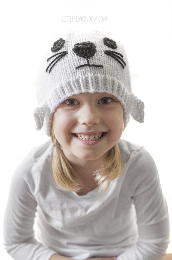 smiling girl in white shirt with blonde braids wearing a white knit hat with a seal face on the front