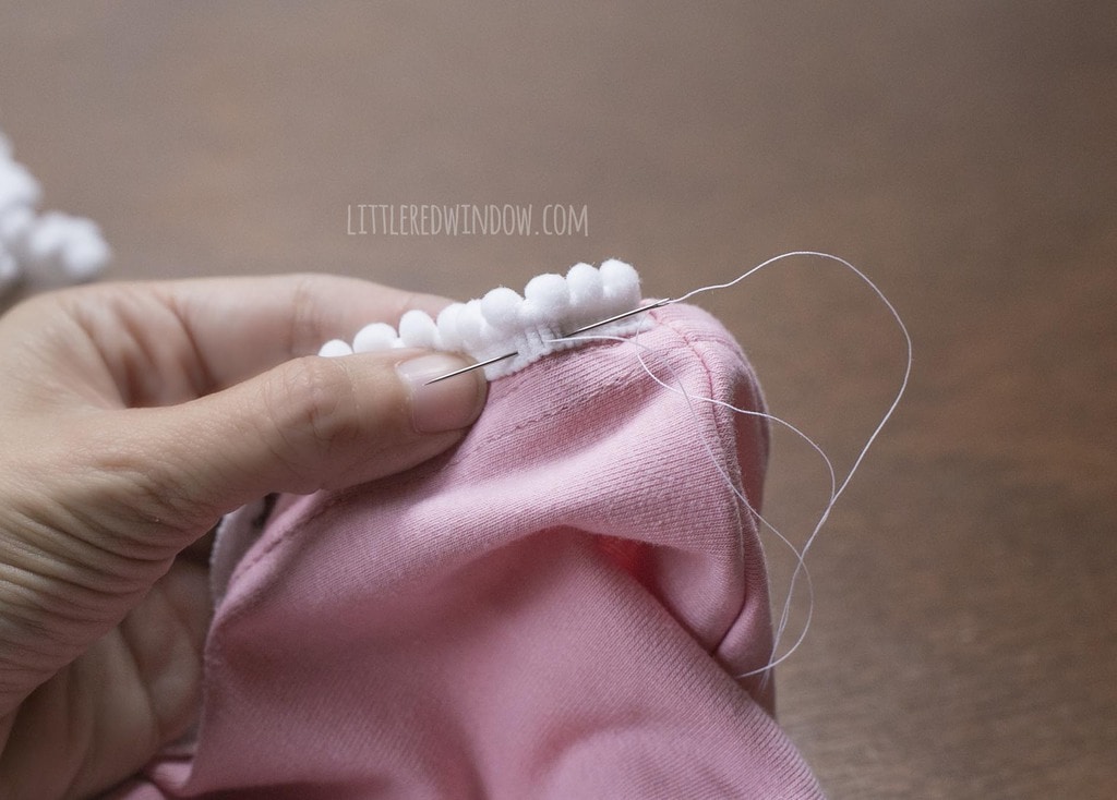 hand sewing white pom pom trim to pink dress hem with white thread and needle