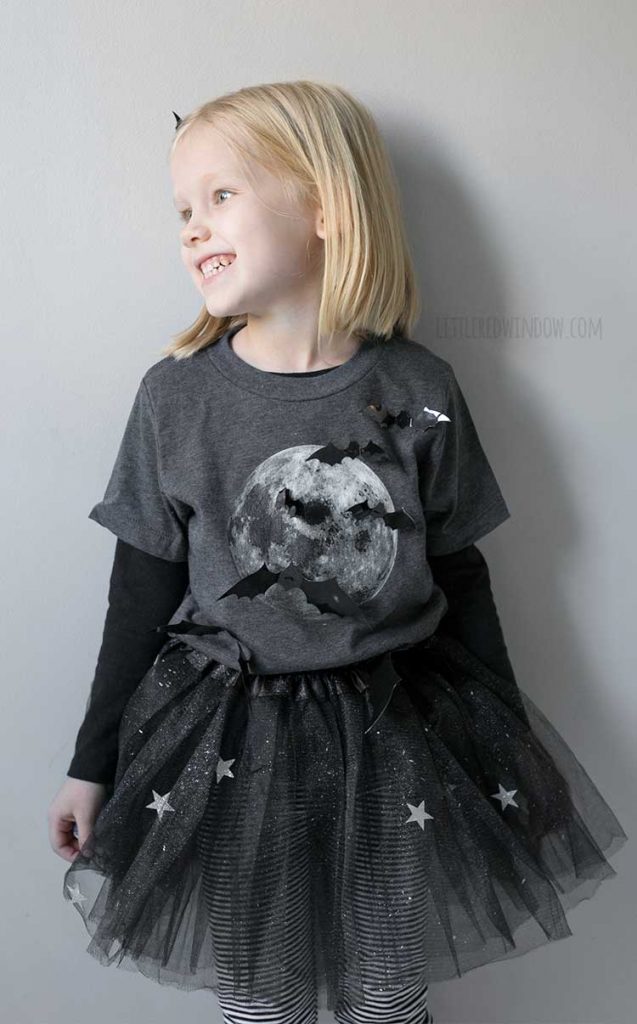 smiling girl looking off to the left wearing bat girl costume with 3D bats over a shirt with a full moon on it and sparking stars tutu