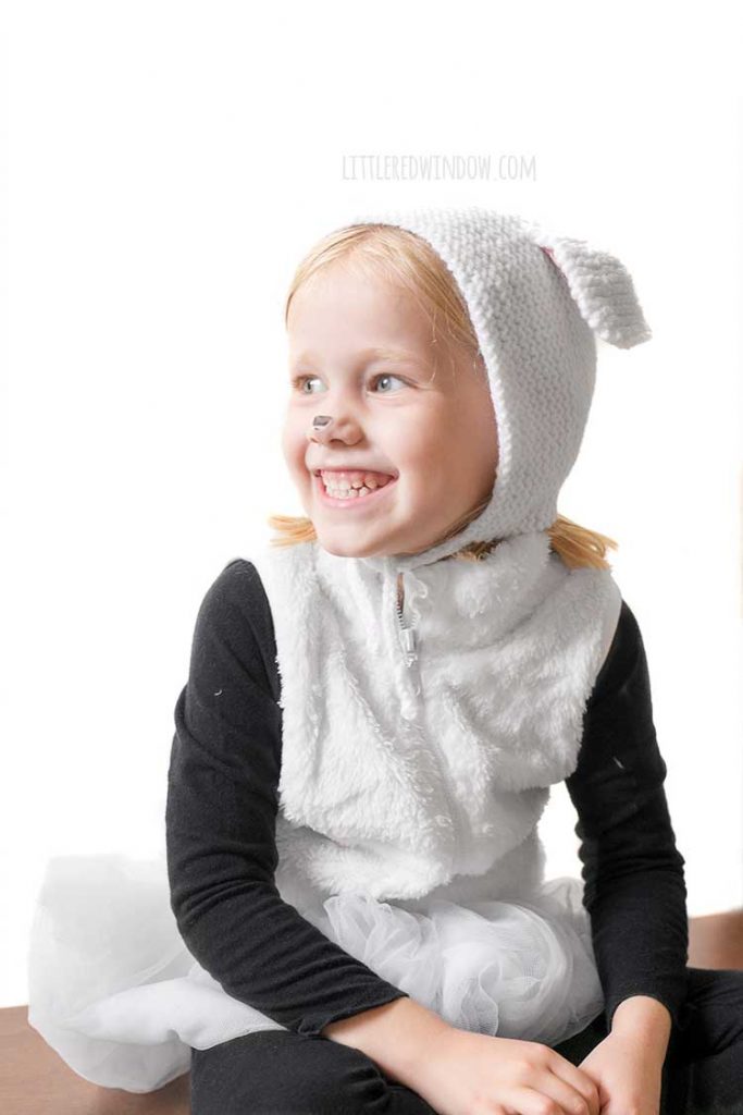 little girl smiling and wearing a white and black sheep costume with sheep ears bonnet looking off to the left over her shoulder