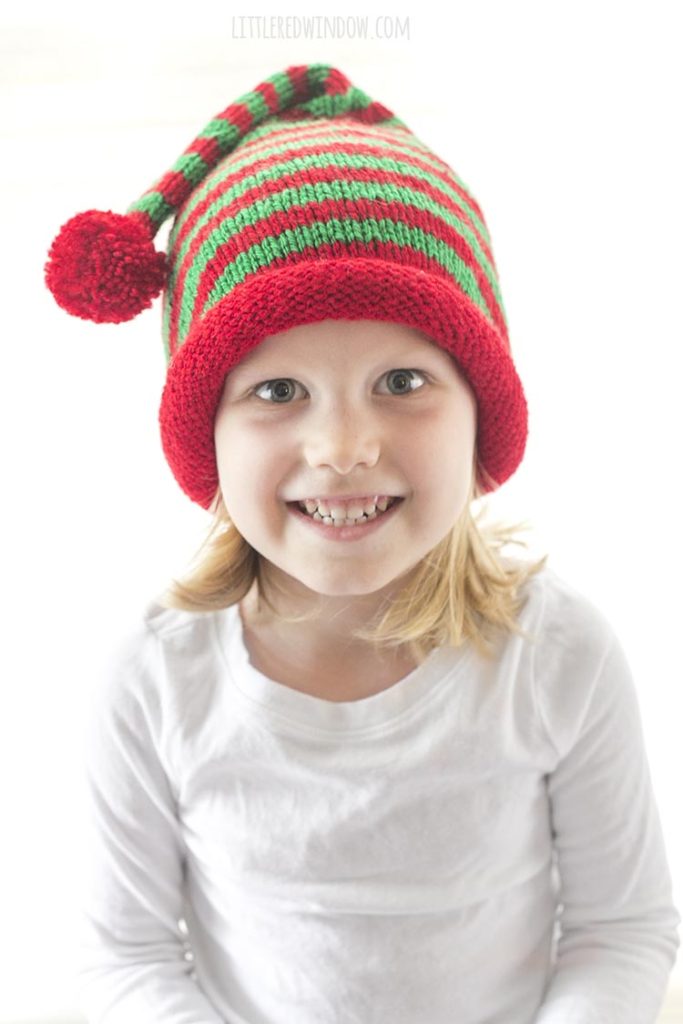 smiling girl looking at the camera and wearing a red and green striped stocking cap with rolled brim and red pom pom