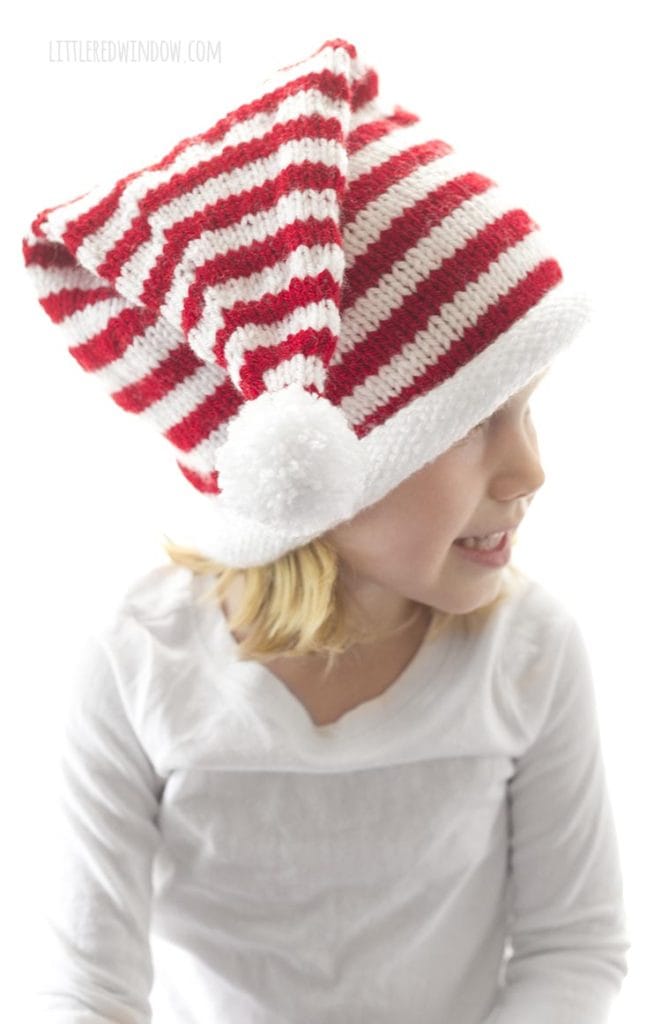 girl in white shirt lwearing a red and white striped knit stocking cap with white rolled brim and white pom pom looking off to the right