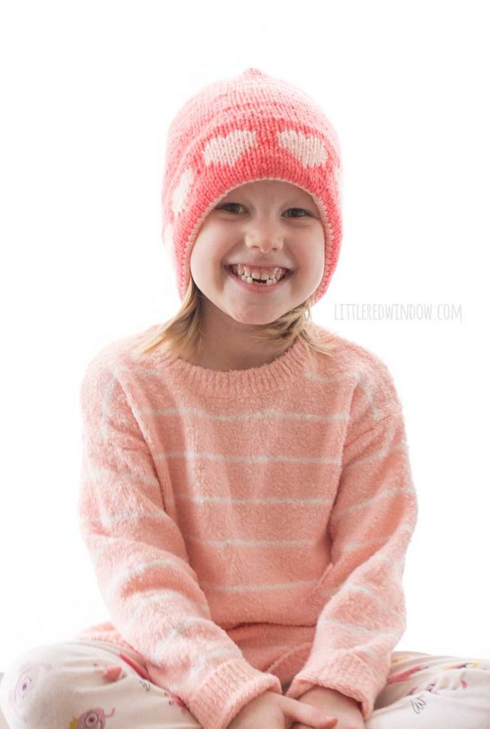 smiling girl in pink sweater wearing pink knit hat with light pink hearts around the brim