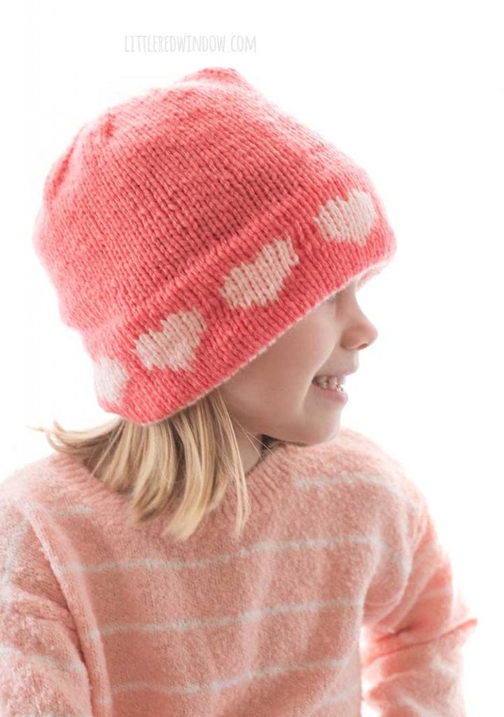 side view of little girl in pink sweater wearing a pink knit hat with light pink hearts around the brim looking off to the right