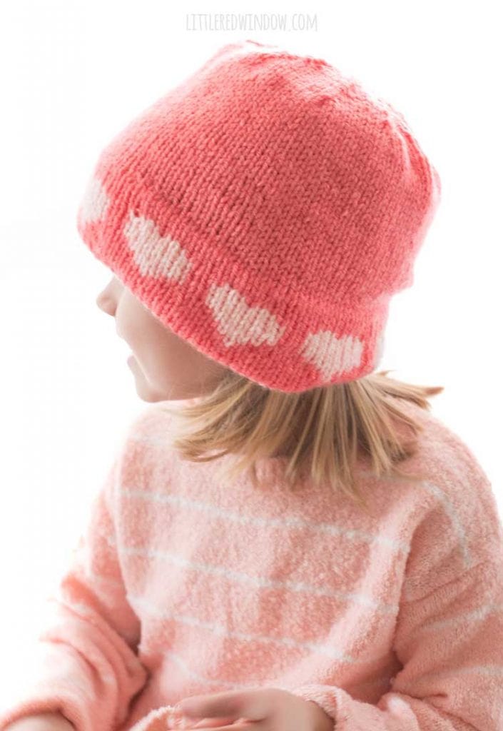 side view of little girl in pink sweater wearing a pink knit hat with light pink hearts around the brim looking off to the left