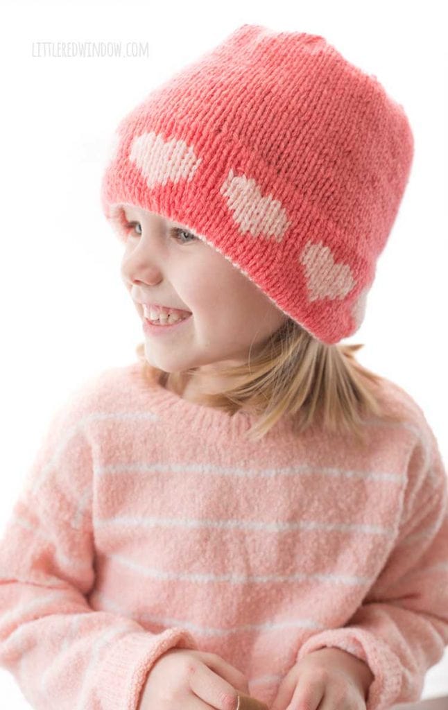 smiling girl in knit pink double brim heart hat looking off to her right