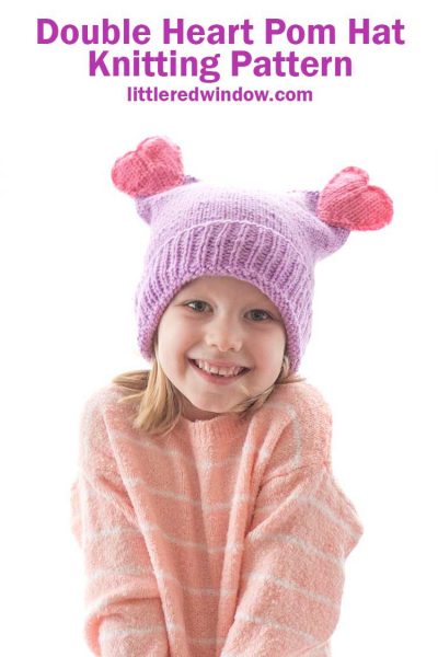 smiling girl in pink sweater wearing a purple knit hat with two pink knit hearts on top