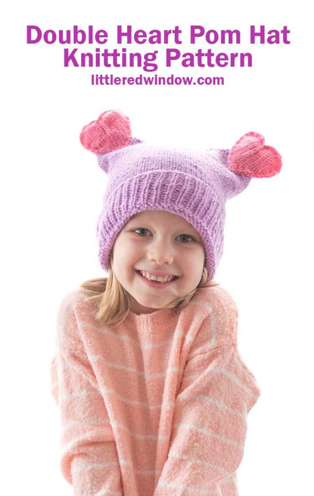 smiling girl in pink sweater wearing a purple knit hat with two pink knit hearts on top