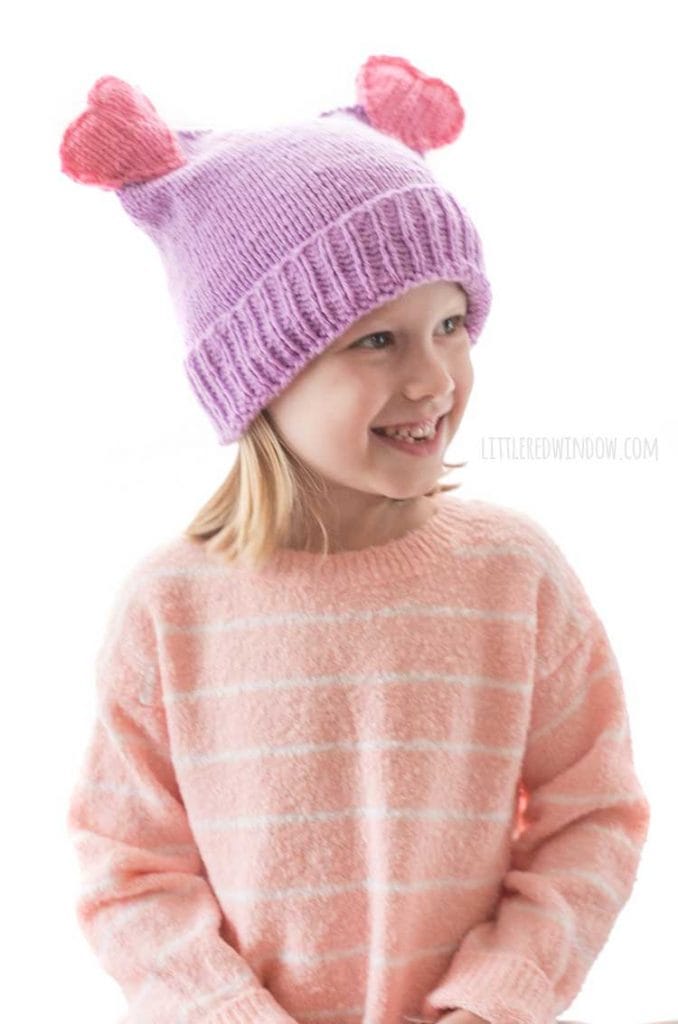 smiling girl looking off in the distance wearing purple and pink double heart pom hat