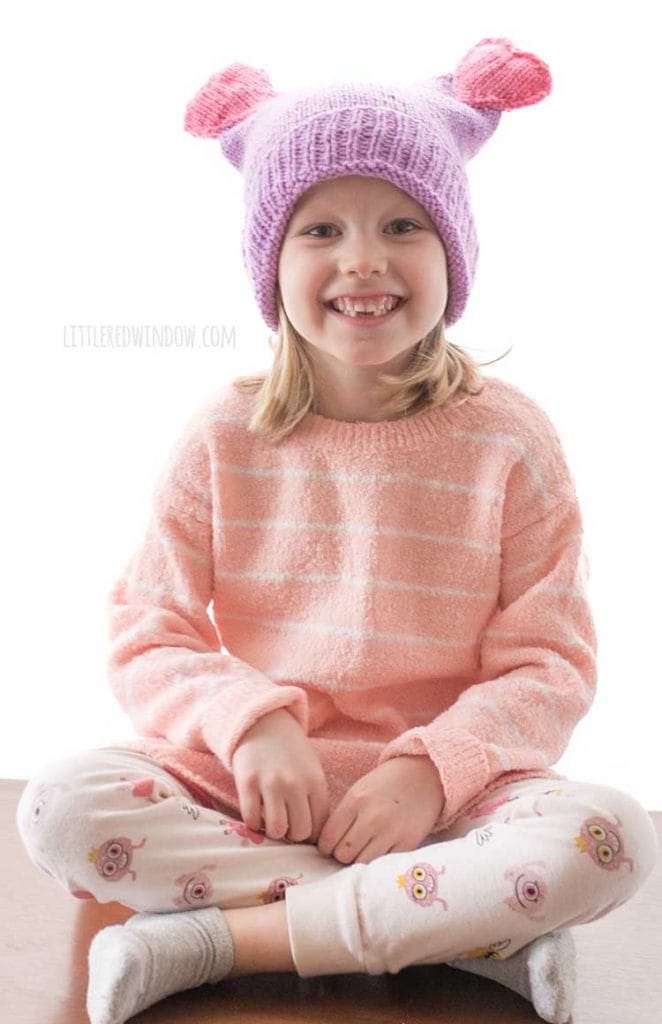 smiling girl sitting cross legged and wearing purple knit hat with double heart poms on the top
