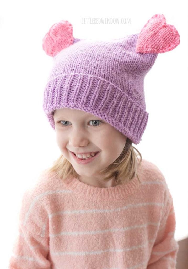 view from above of smiling girl wearing purple and pink double heart pom hat knitting pattern
