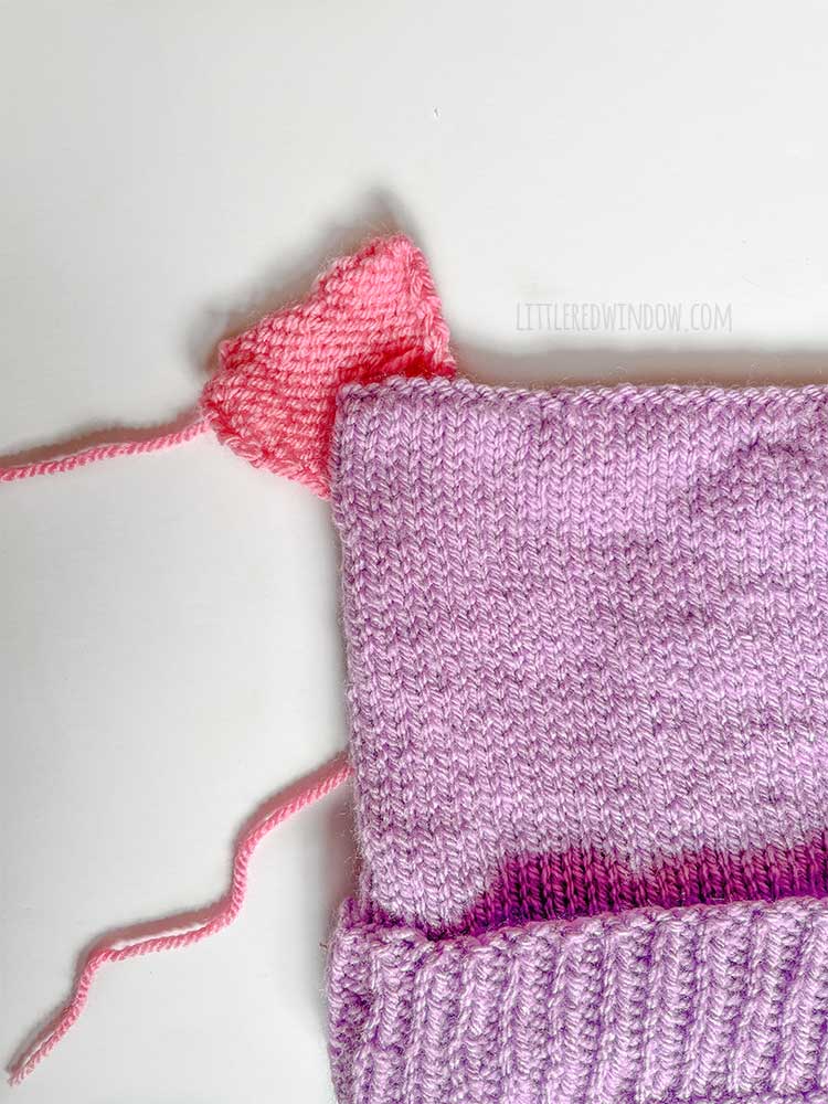 top view of purple knit hat laid out on white background with one knit pink heart placed at the top corner
