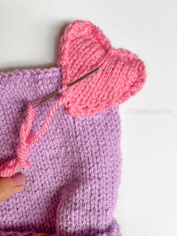 hand sewing the pink hearts to the top of the purple knit hat