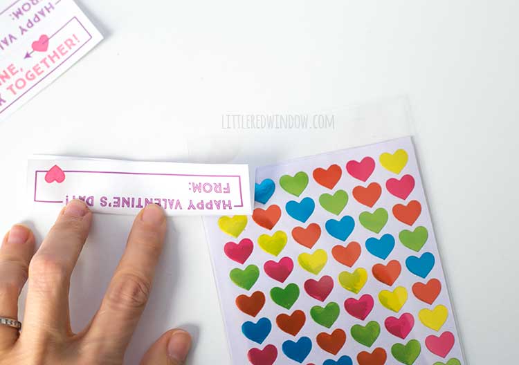 hand folding one printable sticker valentine label next to a sheet of multicolored heart stickers