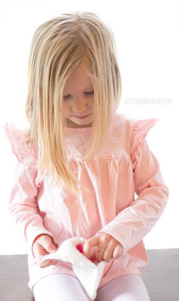 little girl with blond hair and pink shirt pulling red heart our of white knit love letter envelope
