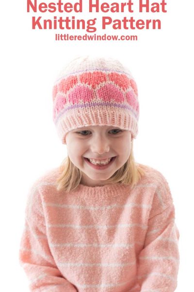 smiling girl in pink sweater wearing a pink knit hat with multicolored pink knit hearts around the middle