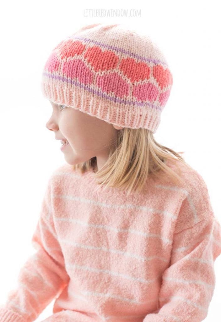 side view of girl in pink sweater wearing a knit pink hat with dark pink and coral nested hearts around the brim looking off to the left
