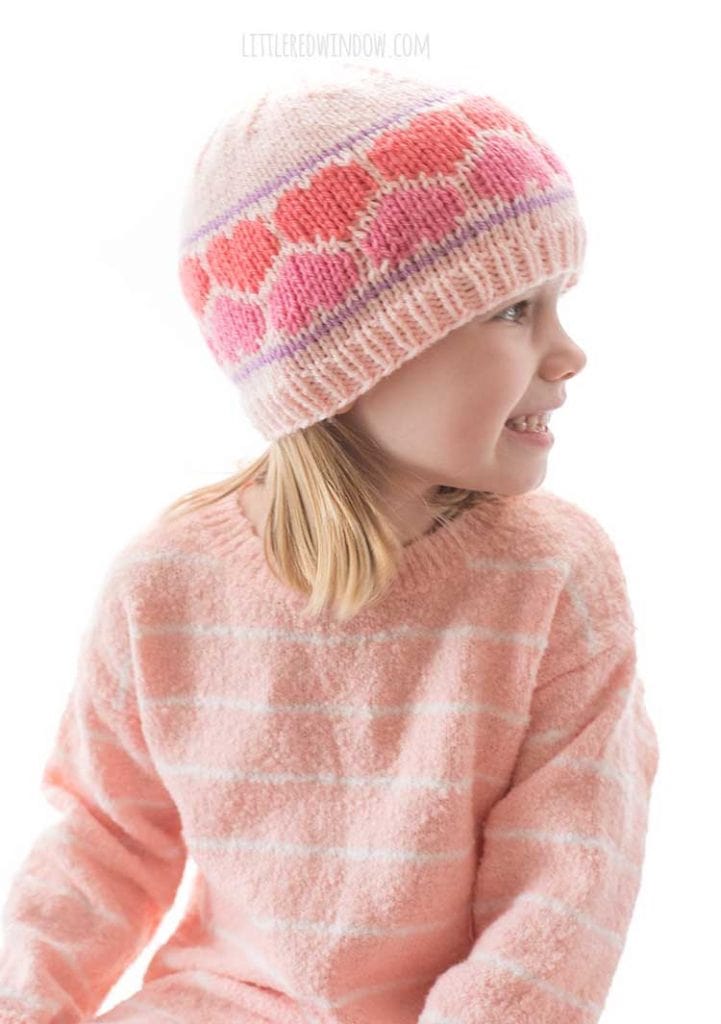side view of girl in pink sweater wearing a knit pink hat with dark pink and coral nested hearts around the brim looking off to the right