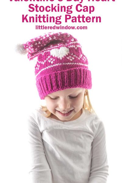 little girl in white shirt wearing magenta colored knit stocking cap with white hearts and white dots on it