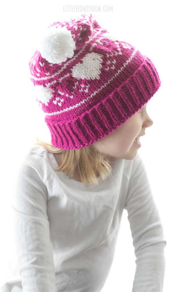 little girl wearing magenta and white knit stocking cap with hearts and polka dots pattern looking off to the right