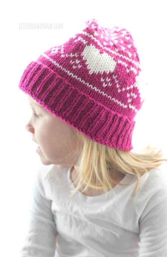 little girl wearing magenta and white knit stocking cap with hearts and polka dots pattern looking off to the left