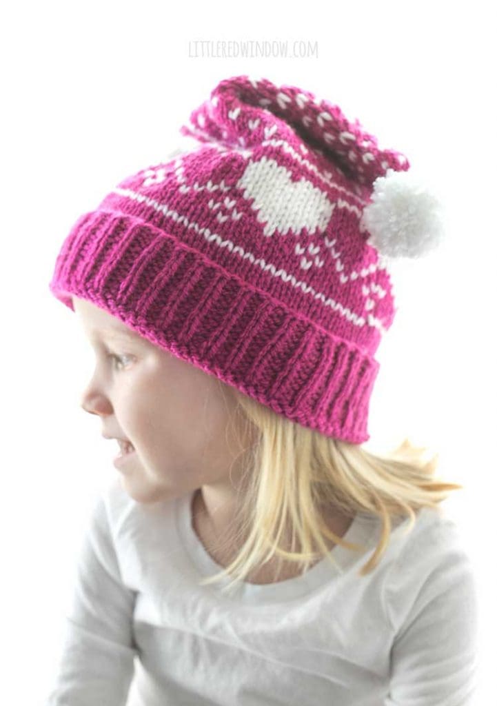 little girl wearing magenta and white knit stocking cap with hearts and polka dots pattern with white pom pom on the end looking off to the left