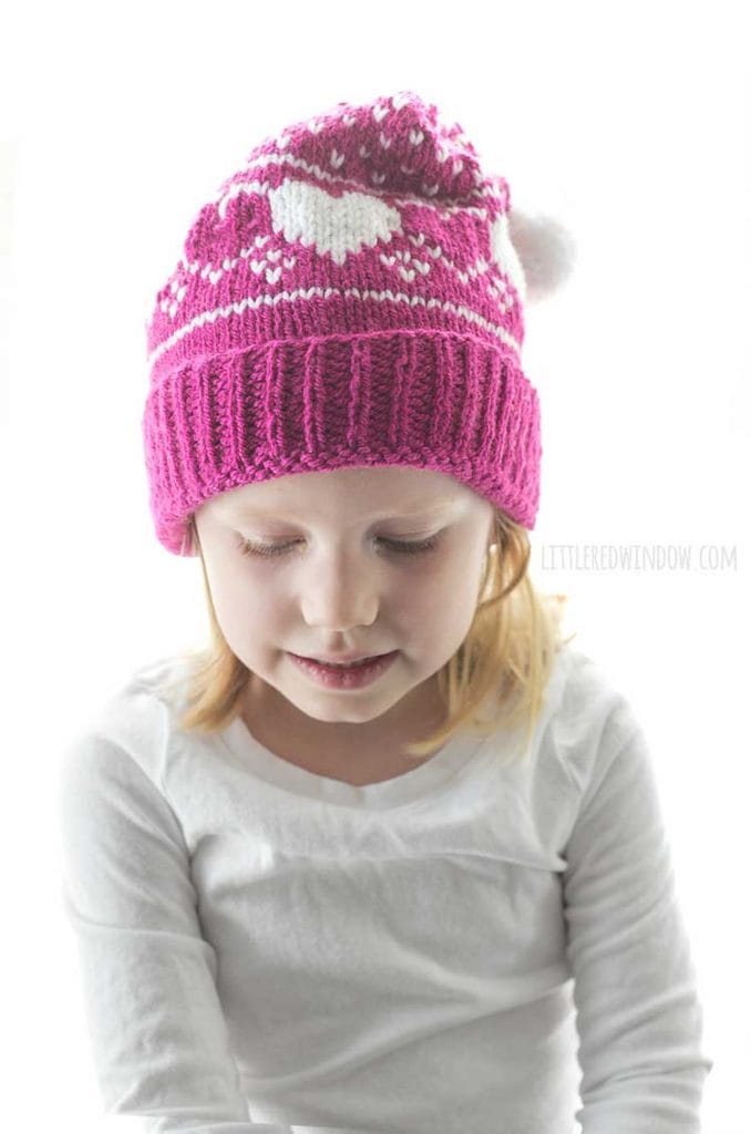 little girl in magenta stocking cap with heart pattern and white pom pom looking down at her lap