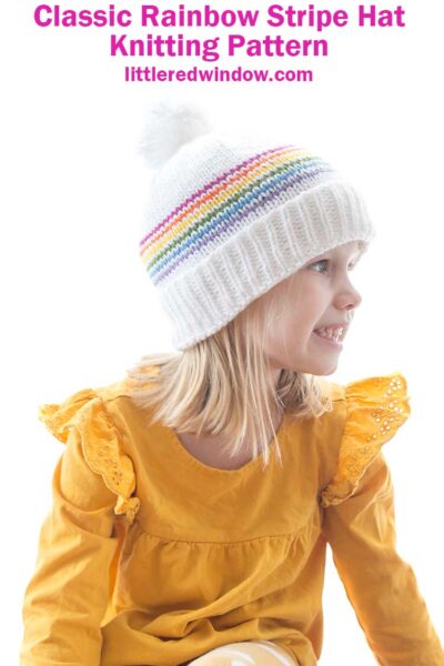 smiling girl in yellow shirt wearing white knit hat with folded brim six thin rainbow stripes and a white pom pom on top looking off to the right