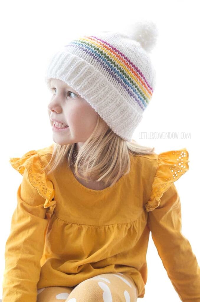 smiling girl in yellow shirt wearing white knit hat with folded brim six thin rainbow stripes and a white pom pom on top looking off to the left