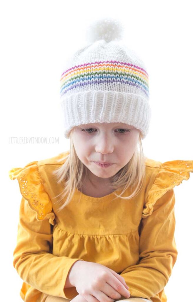 front view of girl in yellow shirt with arms crossed wearing white knit hat with six thin rainbow stripes and white pom pom on top