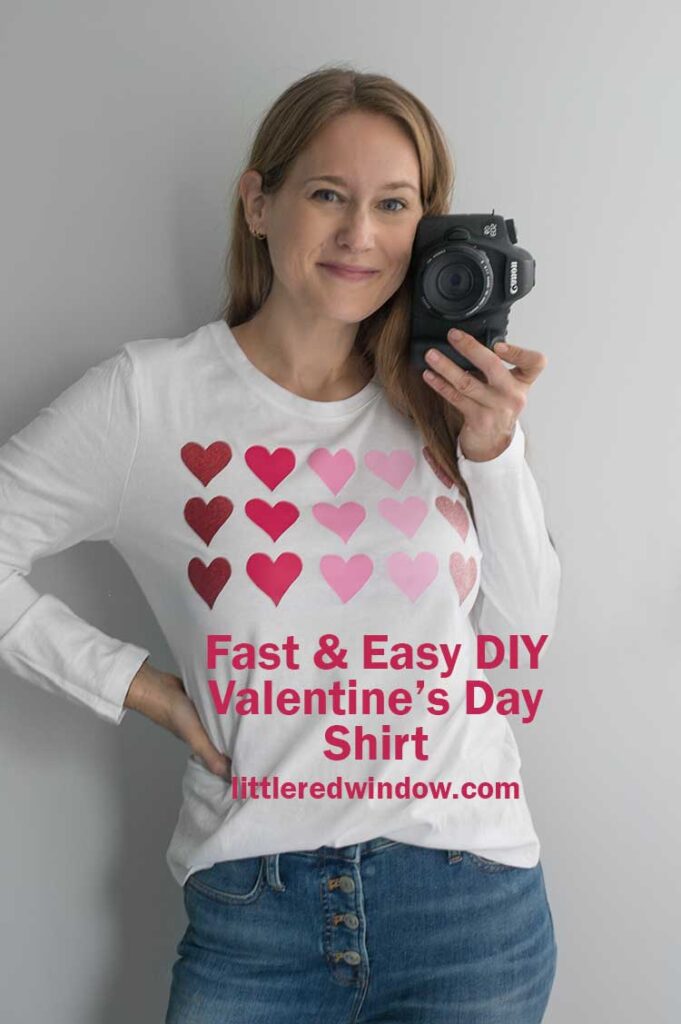 smiling woman holding a camera and wearing jeans and a white long sleeve shirt with pink hearts on the front