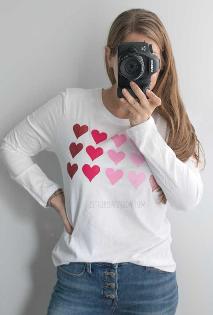 woman wearing jeans and white diy valentines day shirt with 18 pink hearts on the front and holding a dlsr camera in front of her face