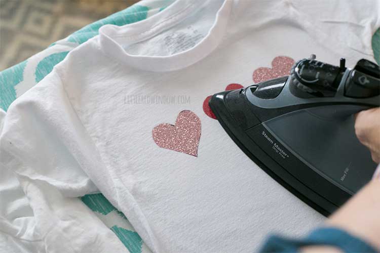 top view of an iron ironing on the glitter heart shapes to the white child tshirt