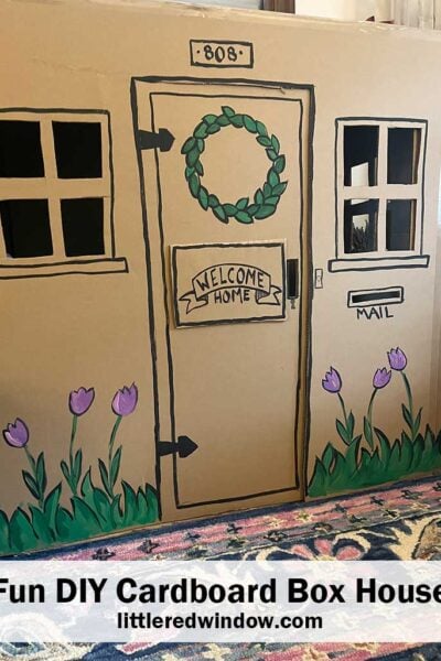 large toy house made out of a cardboard box with a door two windows and flowers painted on the front
