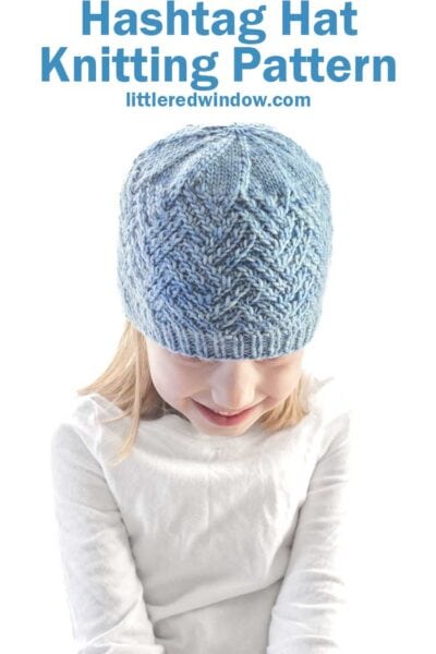 A child in a blue knitted hashtag hat sits against a white background. Above, the text reads "Hashtag Hat Knitting Pattern littleredwindow.com." The child in a white shirt smiles with eyes closed, capturing a moment of cozy joy.