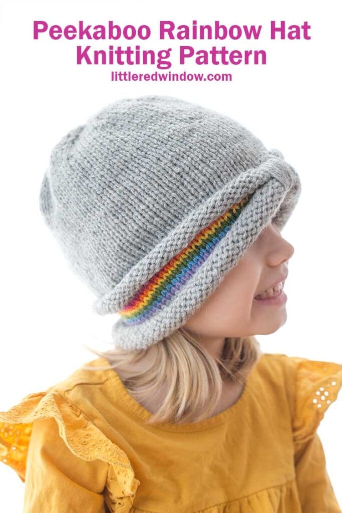 side view of girl in yellow shirt wearing a gray knit hat with a peek of rainbow stripes around the sides