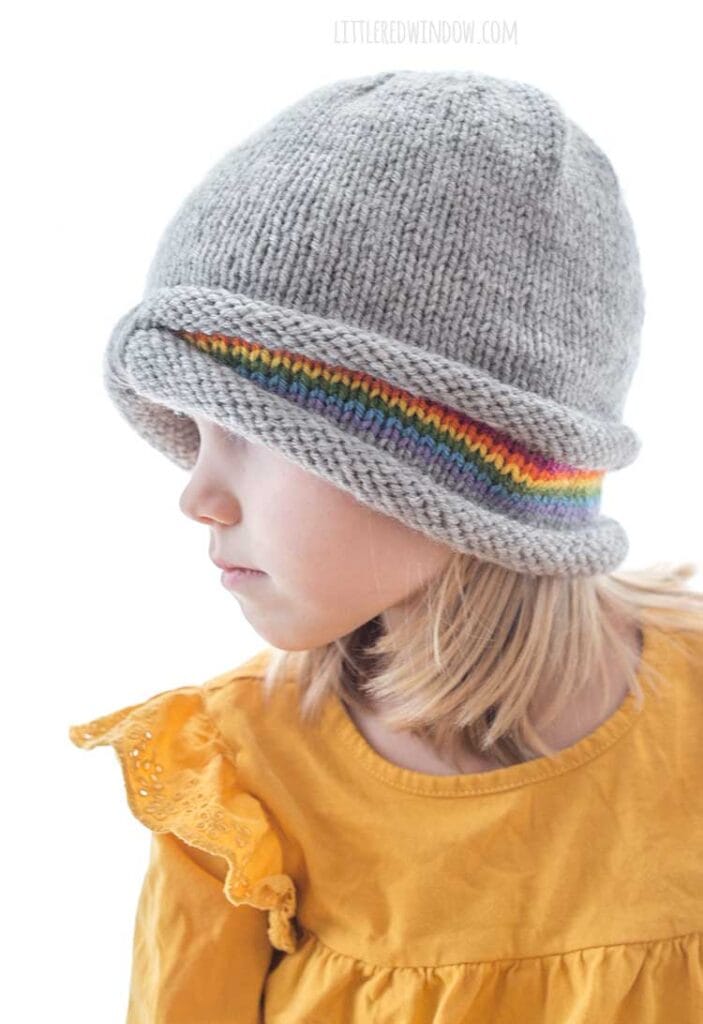 girl in gray knit peekaboo rainbow hat wearing a golden yellow shirt and looking off to the left