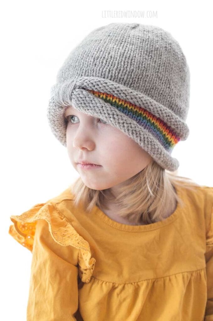 girl in gray knit peekaboo rainbow hat wearing a golden yellow shirt and looking off to the middle left