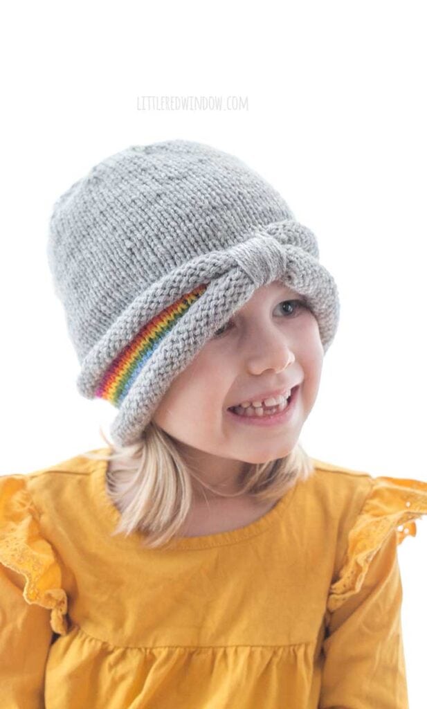smiling girl wearing gray knit hat with gathered brim and peeks of rainbow stripes around the sides