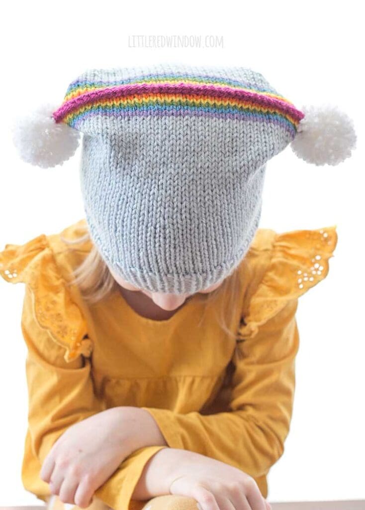 little girl in rainbow double pom hat looking down to show the rainbow and two pom poms on the top