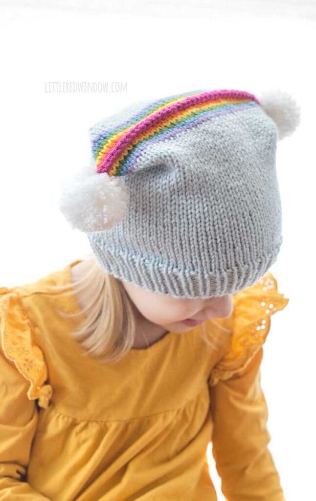 little girl in rainbow double pom hat looking down and to the right to show the rainbow and two pom poms on the top